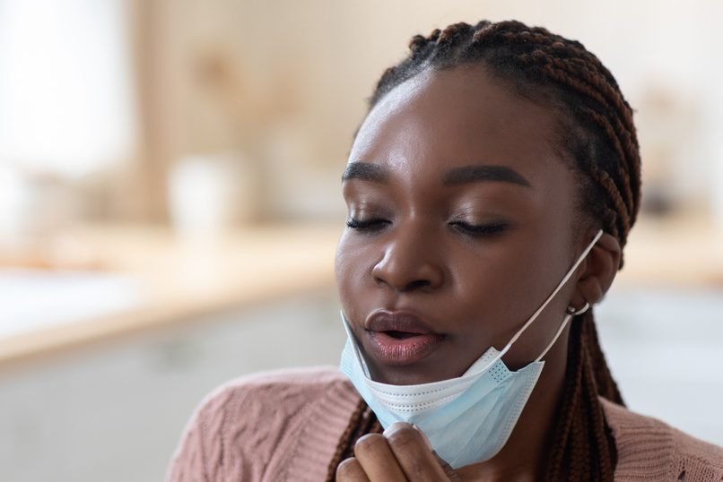 Young Black Woman Taking Off Protective Medical Mask, Suffering Breathing Problems Or Allergy, Making Inhale And Exhale, Having Symptoms Of Coronavirus, Sitting In Kitchen At Home, Closeup Shot. Breathing for health.